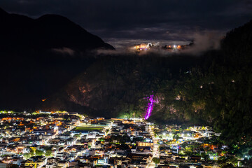 Banos de Agua Santa town center glows at night in Ecuador. Panoramic view features illuminated Cascada de la Virgen waterfall on cliffside above city streets