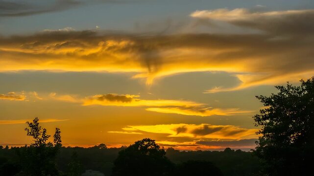 Dramatic funnel cloud formation descends from vibrant orange and yellow sunset clouds over a dark silhouetted horizon of trees