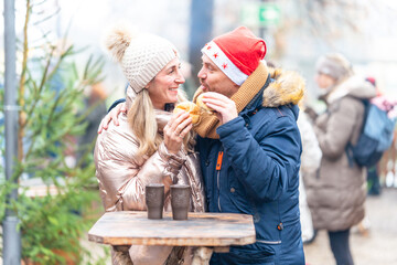 Smiling couple enjoying sausages and rolls at a Christmas market filled with winter warmth and festive charm
