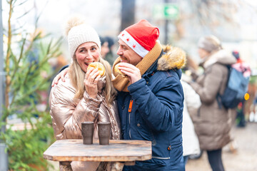 Cheerful couple sharing warm sausages and rolls at a Christmas market enjoying bright holiday lights and flavor