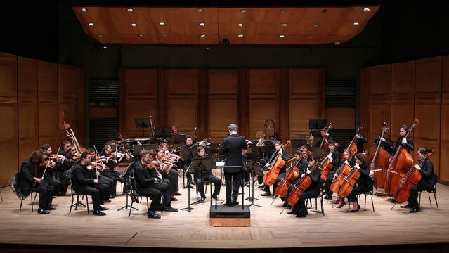 A large group of people in a marching band perform on the city street during a crowded urban parade event while many others walk through the hall