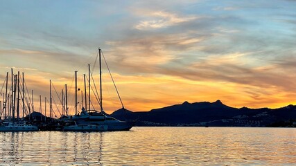 A vibrant sunset over the sea, silhouetting sailboats and yachts docked in a marina, with dark mountains visible across the calm, reflecting water.