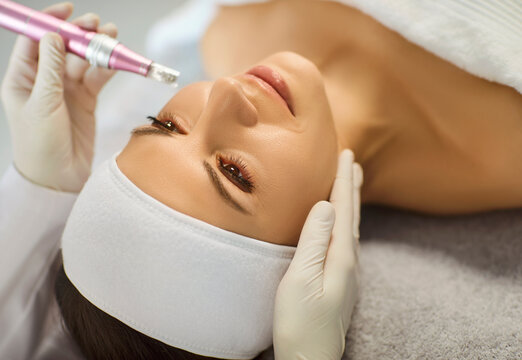 Young woman relaxing during cosmetic microneedling treatment at modern beauty clinic. Cosmetologist using electric dermapen tool on female client face while performing rejuvenating skin care procedure