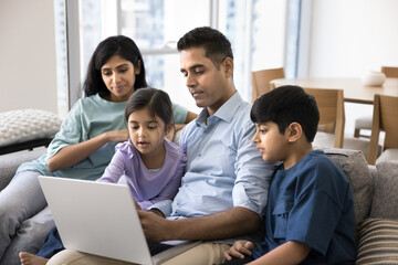 Indian couple with two little kids using laptop at home, choosing goods, making order, purchasing gifts and services remotely through marketplace or e-commerce websites. Tech, family leisure online