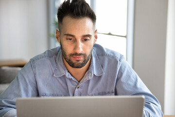Close up Hispanic man sitting at table in homeoffice, focused on his laptop screen with serious expression, working remotely, reading important emails, studying for exam or online course, do research