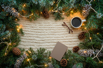 A cozy Christmas frame of evergreen boughs, pinecones, lit strings, a mug, and glasses on a knitted background. Ideal for holiday reading, winter aesthetic, and cozy moments concepts. copy space