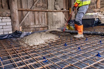 A groundworker skillfully pours concrete onto a metal grid at a construction site. This image...