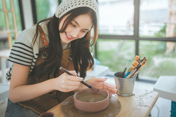 Asian woman artist painting handmade ceramic product in pottery studio – creative process of decorating sculpture with brush for small business, art workshop, and modern craft design startup