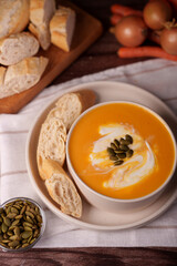 warm and inviting autumnal meal setup with pumpkin soup garnished with cream and pumpkin seeds, accompanied by sliced rustic bread and fresh vegetables on a wooden and cloth surface, setup, serving