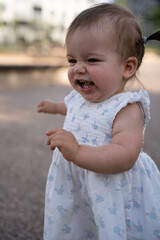 joyful toddler in white floral dress laughing and playing outside on a sunny day in a blurred urban background with soft natural light and warm atmosphere, delight, childhood, carefree, spontaneous