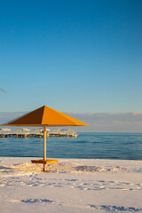 A single yellow sun umbrella stands on a snow-covered beach with a long resort pier visible in the distance across the blue waters of Issyk-Kul Lake