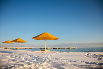 Three bright yellow sun umbrellas stand on a snow-covered beach overlooking a long resort pier on the blue waters of Issyk-Kul Lake