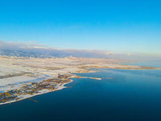 Wide aerial view of a large body of deep blue water meeting a vast, snow-covered landscape with scattered buildings and distant snow-capped mountains