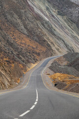 An asphalt road winds through arid mountainsides revealing layers of multi-colored sedimentary rock and patches of dry grass