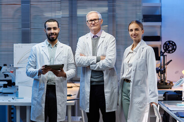 Group of three scientists standing in laboratory, aged Caucasian man with arms crossed, young adult Caucasian woman and young adult man holding digital tablet, all wearing lab coats