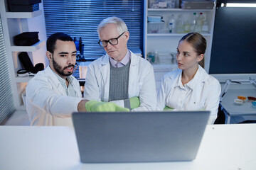 Middle aged man, senior Caucasian man, young adult Caucasian woman wearing lab coats collaborating and analyzing data on laptop in laboratory setting, focused on screen