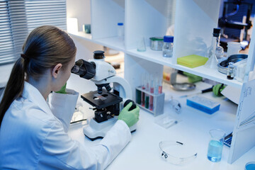 Caucasian young adult woman examining samples using microscope in laboratory setting, wearing lab coat and gloves, surrounded by scientific equipment and glassware on workbench