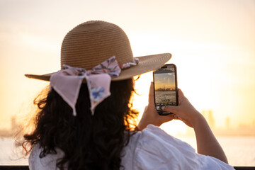 Tourist woman photographing Dubai skyline at sunset with smartphone capturing golden hour waterfront view and iconic Burj Khalifa cityscape for travel lifestyle and tourism concept © Nirja