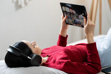 Young Asian woman relaxing on a sofa, wearing headphones, and using a digital tablet for entertainment or browsing.