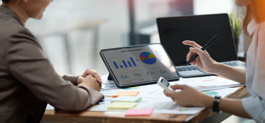 Two female professionals analyze a digital business report on a tablet, working collaboratively on market trends and strategy in a bright modern office setting.