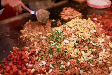 Cook preparing savory street food meal on large grill. Delicious food featuring mix of meat, vegetable, and egg stirred with spatula during cooking. vibrant and appetizing dish