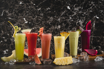 Six vibrant fruit juices and smoothies with fresh fruits and ice cubes on a dark marble background.