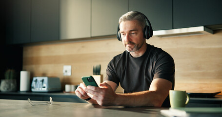 Headphones, streaming or man in home with phone, song connectivity or listening to music playlist. Sound track, mature or male person in kitchen with tech, podcast or radio subscription on audio app.