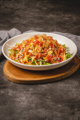 A vibrant and fresh Asian-style salad or noodle dish with green vegetables, yellow strips, red sauce, and crispy toppings served in a white bowl on a wooden board.