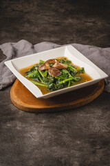 A square white plate of stir-fried green leafy vegetables with mushrooms in a savory sauce, served on a round wooden board.