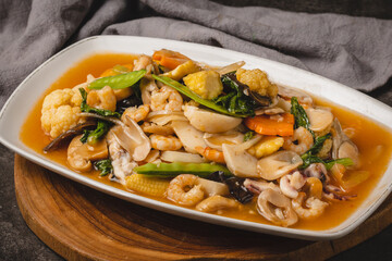A vibrant plate of stir-fried mixed seafood and vegetables, including shrimp, squid, mushrooms, carrots, and snow peas, served in a savory sauce on a white rectangular dish.