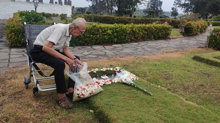 A lonely, elderly man in a wheelchair, mourning a loved one, tenderly places flowers on a grave or memorial in a peaceful cemetery park. Symbolizes loss, remembrance, grief, old age, and enduring love