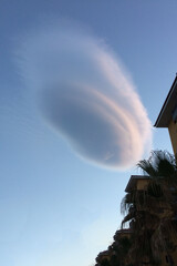 A unique, elongated cloud formation floats in a clear blue sky above the rooftops of a coastal building. The unusual shape of the cloud for a travel reel.