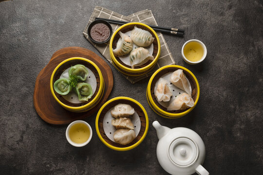 An overhead shot of a delicious assortment of steamed dim sum dumplings in bamboo baskets, served with hot tea on a dark table.
