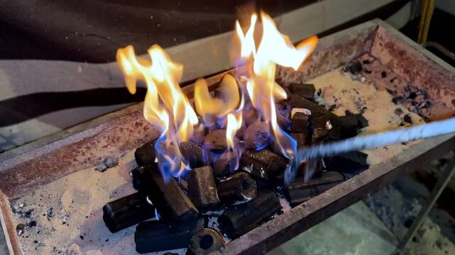 Close-up of metal tongs arranging glowing hexagonal charcoal briquettes in a sand-lined grill. Blue and orange flames rise from the hot coals, creating intense heat for outdoor cooking or BBQ.