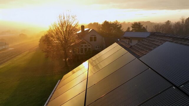 Golden sunset light reflects on rooftop solar panels in quiet American suburban neighborhood, with misty autumn trees and modern family home. Aerial close up backwards shot in suburbia of US.