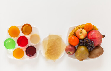 Top view of colorful fruit jelly, gelatin sheets, and plate of vitamin fruits for making delicious and healthy dietary treat. White background, empty space for recipe, flat lay, closeup, copy space