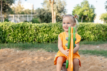 A young Caucasian girl with blonde hair sits on a playground seesaw