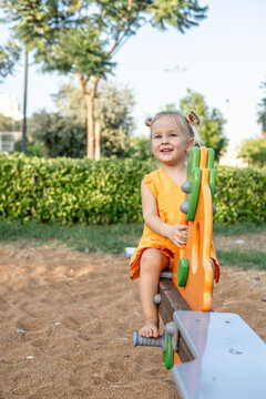 young girl with blonde hair kghappily sits on playground seesaw