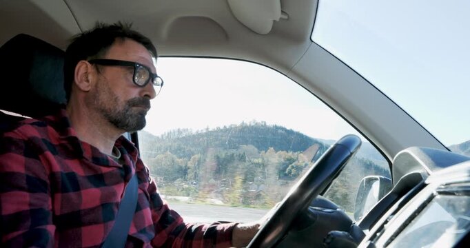 A driver navigating through a rural town is seen from the side, passing buildings and hills on a sunny day.
