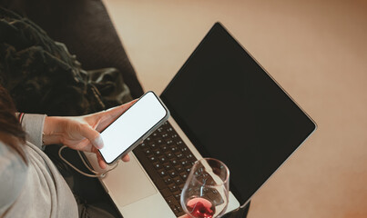 Woman holding smartphone with blank screen and drinking wine beside laptop, representing digital lifestyle,Businesswoman relaxation after work, social media browsing, or modern multitasking at home