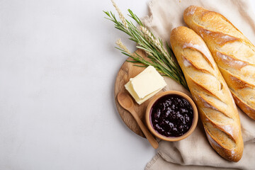 Freshly baked artisan bread and slices with butter, along with a bowl of strawberry jam, placed on a light linen cloth, surrounded by wheat ears and rosemary
