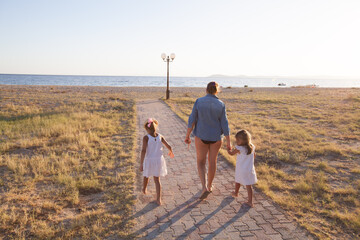 Family walking hand-in-hand toward beach along sunlit path with sea and boats