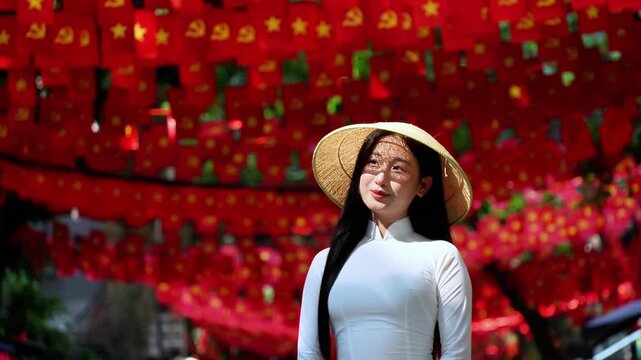 A young Vietnamese woman wearing a traditional white ao dai and a conical hat smiles gracefully under bright sunlight, in front of vibrant red Vietnam flags decorations