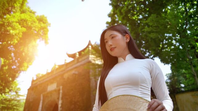 A young Vietnamese woman wearing a traditional white ao dai and a conical hat smiles gracefully under bright sunlight, on street in Hanoi, Vietnam.