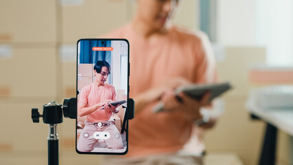 Close up youth Asia male owner small business online shop sit front of phone look at camera live streaming see comment on digital tablet from customer sales promotion at retail online shop at home.