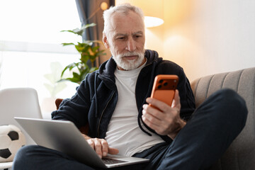 Senior man relaxing on sofa, using smartphone and laptop, enjoying technology at home