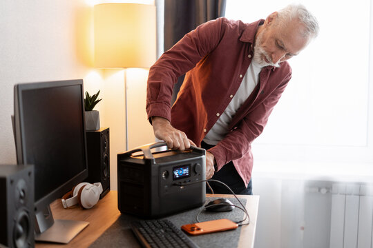 Senior man plugging smart device into portable power station at home office desk
