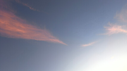 Two wispy long clouds with bright peach and rose-gold edges stretch across clear blue-grey sky...