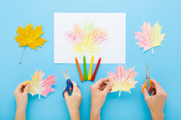 Mother and little child hands cutting paper leaves on blue table background. Pastel color. Painting and copying colorful maple leaf pattern. Closeup. Making autumn decoration. Flat lay. Top down view.