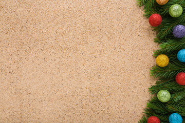 Colorful baubles with glitter and green natural fresh fir branches on light brown dry sea sand background. Closeup. Christmas decoration. Empty place for text. Top down view.